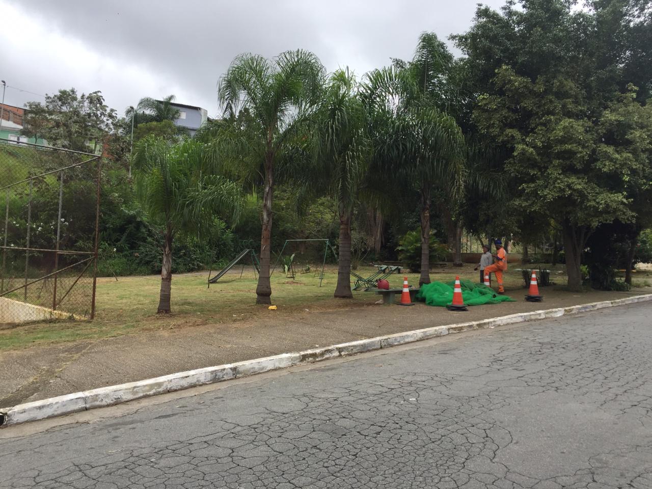 Na beira de uma parte de alambrado, indicando uma quadra de esportes, a grama foi cortada, três coqueiros se destacam na paisagem. Um trabalhador, com uniforme laranja, conversa com um homem de chapéu, vestido com uma camisa branca  e calça escura. Na frente deles, a tela de proteção verde já está enrolada e ao lado, quatro cones de sinalização. 